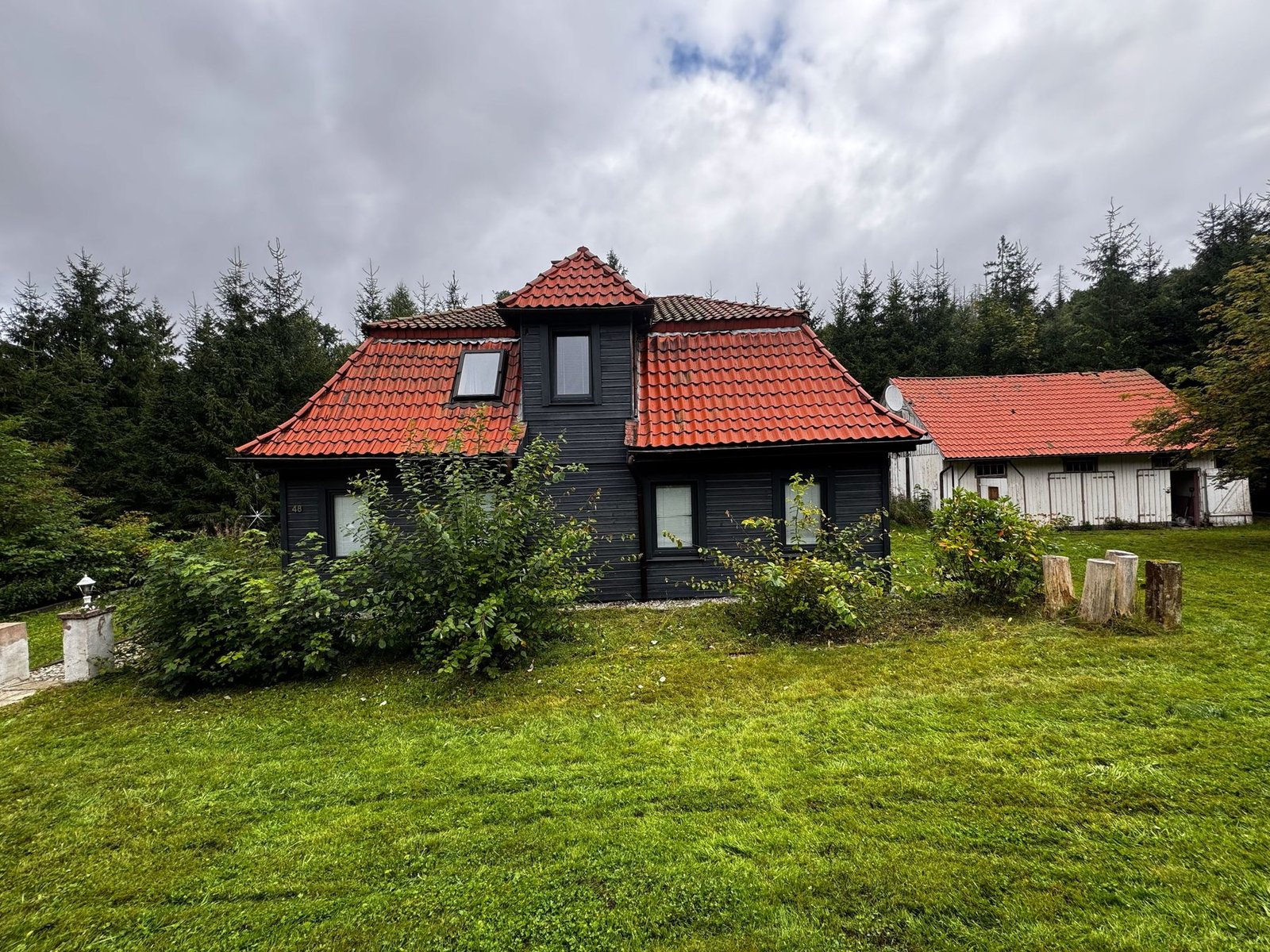 Das dunkel getäfelte Ferienhaus mit rotem Ziegeldach zwischen Wald und Nebengebäude.