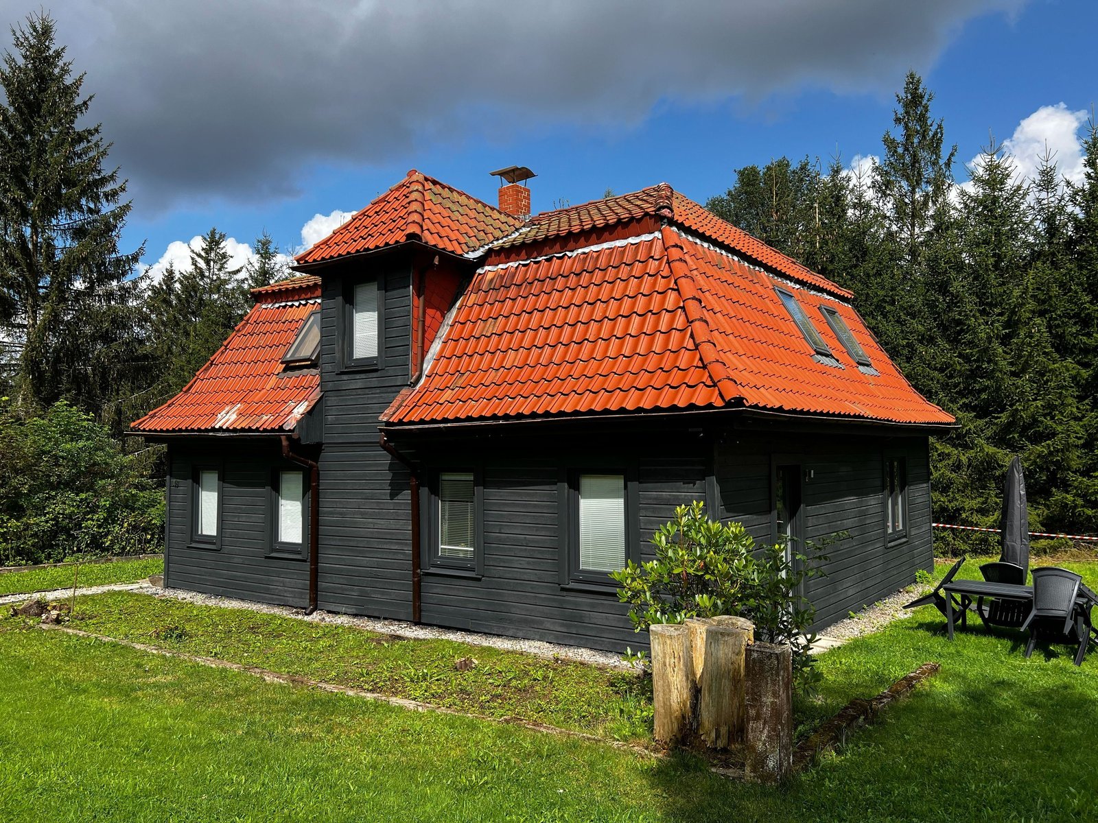 Das Ferienhaus am Waldschwimmbad mit dunkler Holzfassade und rotem Ziegeldach unter dramatischem Himmel, umgeben von Wald.